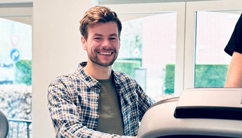 Man met een geruite blouse glimlacht terwijl hij op een loopband staat.