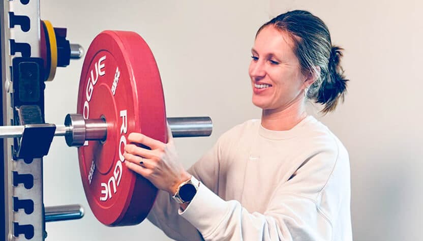 Vrouw in een crèmekleurige trui plaatst een gewicht op een halter in een fitnessruimte.
